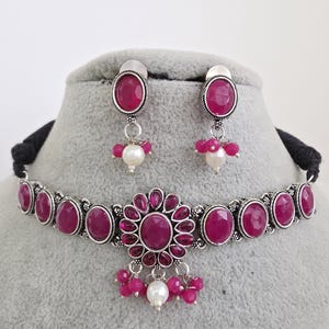 May include: A silver-toned choker necklace and matching earrings set. The necklace features oval, magenta-colored stones set in a floral design. The earrings and necklace have a black cord. Each earring has a magenta bead cluster and a white pearl.