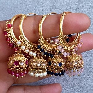 May include: Four gold-colored hoop earrings with dangling bell-shaped charms. Each earring features a different color of small beads, including red, white, blue, and pink. The earrings are held up by a hand.