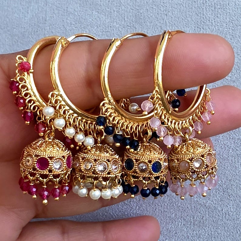 May include: Four gold-colored hoop earrings with dangling bell-shaped charms. Each earring features a different color of small beads, including red, white, blue, and pink. The earrings are held up by a hand.
