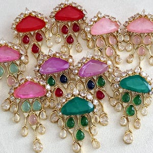 Può includere: Una collezione di orecchini dorati e ornati con accenti di pietre preziose colorate. Ogni orecchino presenta una grande pietra a forma triangolare nei toni del rosso, rosa, viola e verde, circondata da gemme più piccole e cristalli a goccia pendenti.