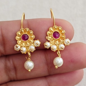 May include: A pair of gold-colored earrings. Each earring features a flower-shaped design with a red center stone and small white pearls. A larger white pearl dangles from the bottom of each earring.