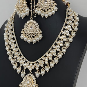 May include: A gold and white Indian bridal necklace set with pearl accents. The set includes a long necklace, a shorter necklace, a pair of earrings, and a maang tikka.