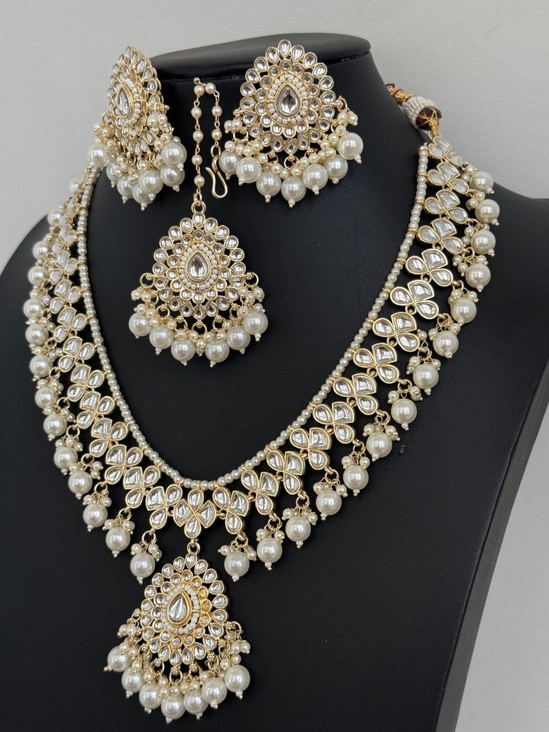 May include: A gold and white Indian bridal necklace set with pearl accents. The set includes a long necklace, a shorter necklace, a pair of earrings, and a maang tikka.