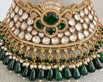 May include: A gold-toned statement necklace featuring emerald green and clear gemstones. The necklace has a wide collar design with a central cluster of green stones and dangling green teardrop-shaped beads. The necklace is adorned with small white pearls.