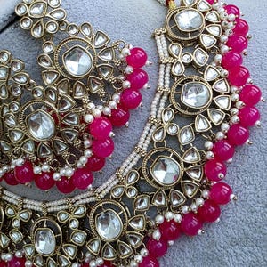 May include: A gold-toned necklace and earring set featuring large, clear, faceted stones set in ornate metal frames. The jewelry is accented with vibrant hot pink spherical beads and small white seed pearls arranged in layered, dangling patterns.