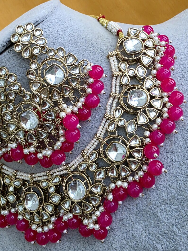 May include: A gold-toned necklace and earring set featuring large, clear, faceted stones set in ornate metal frames. The jewelry is accented with vibrant hot pink spherical beads and small white seed pearls arranged in layered, dangling patterns.