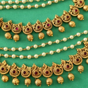 Könnte beinhalten: Nahaufnahme von goldfarbenem Schmuck mit roten Edelsteinakzenten und baumelnden goldenen Perlen. Der Schmuck weist aufwendige, von Pfauen inspirierte Designs auf und ist mit Strängen aus weißen Kunstperlen kombiniert. Der Hintergrund ist einfarbig türkis.