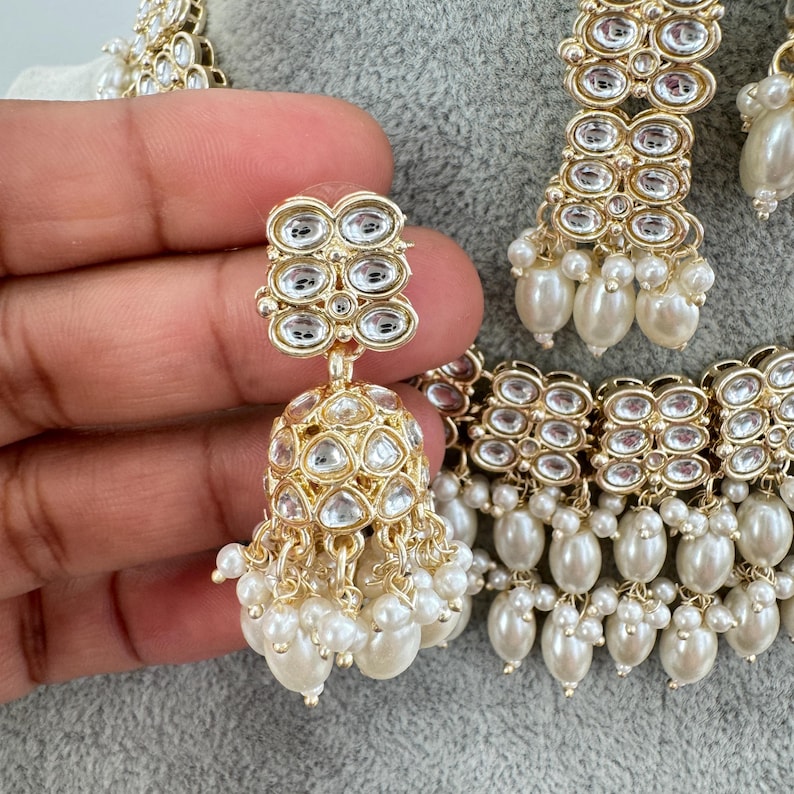 May include: A gold necklace with a multi-layered design featuring clear stones and white pearls. The necklace is paired with matching earrings with a similar design.