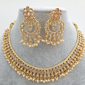 May include: A gold-coloured necklace and earring set displayed on a mannequin. The jewellery features clear oval-shaped stones and small white pearls. The earrings are a half-moon shape. The necklace and earrings are connected by a gold chain.