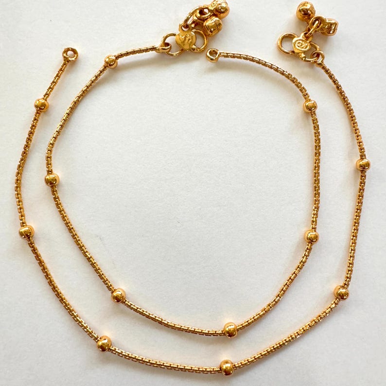 K&ouml;nnte beinhalten: Ein Paar goldene Fu&szlig;kettchen mit kleinen runden Perlen und dekorativen Anh&auml;ngern. Die Fu&szlig;kettchen bestehen aus einer zarten Kette und sind dazu bestimmt, zusammen getragen zu werden.