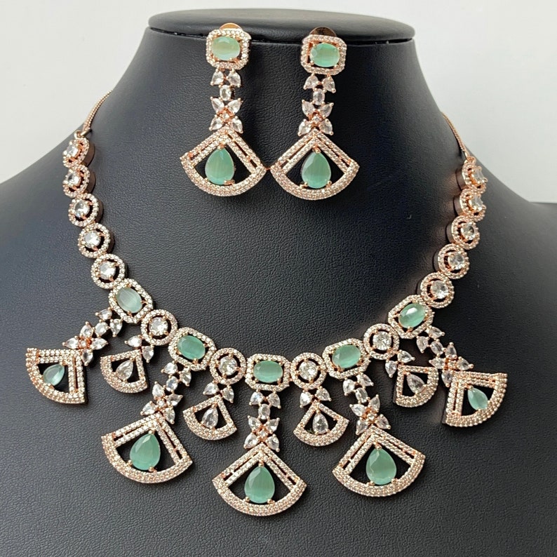 Pode incluir: Um conjunto de colar e brincos de ouro rosa com um design delicado com pedras transparentes e verde claro. O colar tem um pingente em forma de leque com v&aacute;rias camadas.