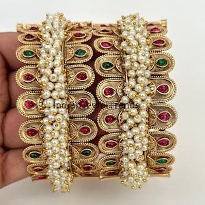 May include: Two gold bangles with red and green gemstones and white pearl accents.