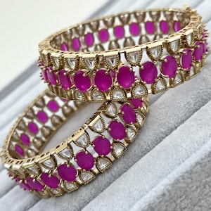 May include: A pair of gold-toned bangles featuring a repeating pattern of oval-shaped vibrant magenta stones alternating with clear triangular stones. The metal framework has a textured, beaded edge design surrounding each individual gemstone setting.