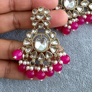 May include: A gold-toned metal earring featuring a central clear stone surrounded by smaller teardrop-shaped stones. The design includes dangling bright pink spherical beads and tiny white seed pearls arranged in a decorative, ornate pattern.