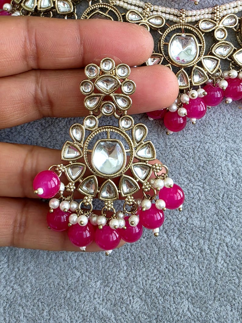 May include: A gold-toned metal earring featuring a central clear stone surrounded by smaller teardrop-shaped stones. The design includes dangling bright pink spherical beads and tiny white seed pearls arranged in a decorative, ornate pattern.