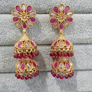 May include: A pair of gold-coloured earrings with a floral design. The earrings feature a flower-shaped stud with pink and clear gemstones. The dangling part has a bell shape with pink gemstones and small red beads.