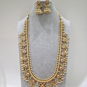 Può includere: Un set di collana e orecchini dorati. La collana presenta un design lungo e ornato con dettagli rossi e bianchi. Gli orecchini sono un gruppo di perle bianche e dettagli rossi. I gioielli sono esposti su un manichino grigio.