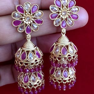 Puede incluir: Un par de pendientes jhumka dorados con un poste en forma de flor con piedras alternas en forma de lágrima de color fucsia y transparente. El colgante en forma de campana tiene dos niveles decorados con piedras y cuentas fucsias.