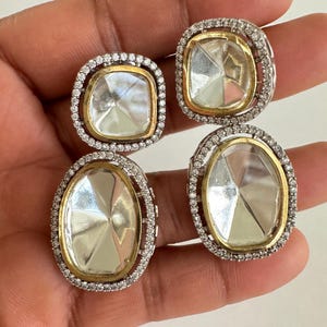 May include: Four square and oval-shaped earrings. Each earring features a clear, faceted center stone set in a gold-toned bezel. The stones are surrounded by a halo of small, clear stones set in a silver-toned metal.