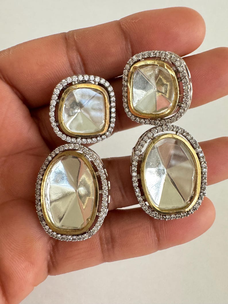 May include: Four square and oval-shaped earrings. Each earring features a clear, faceted center stone set in a gold-toned bezel. The stones are surrounded by a halo of small, clear stones set in a silver-toned metal.