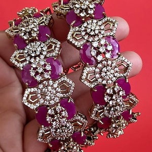 May include: Two gold-tone bangles featuring a repeating geometric pattern. Each bangle is set with vibrant magenta oval-shaped stones and numerous small, clear, sparkling white crystals arranged in intricate floral-inspired clusters.