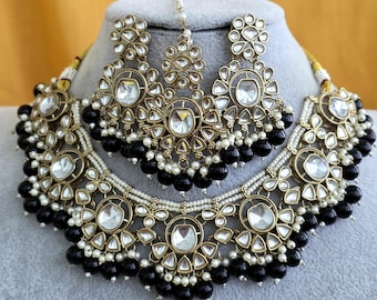 Op de afbeelding: Een goudkleurige sieradenset bestaande uit een ketting, twee oorbellen en een maang tikka. Elk sieraad is versierd met heldere, druppelvormige stenen, kleine witte kraaltjes en grotere ronde zwarte kralen in metalen zettingen.