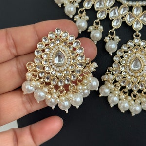May include: A gold and white necklace and earrings set with pearl accents. The necklace features a layered design with multiple tiers of pearls and floral-shaped embellishments. The earrings are similarly designed with a round, floral-shaped center piece and a dangling pearl drop.