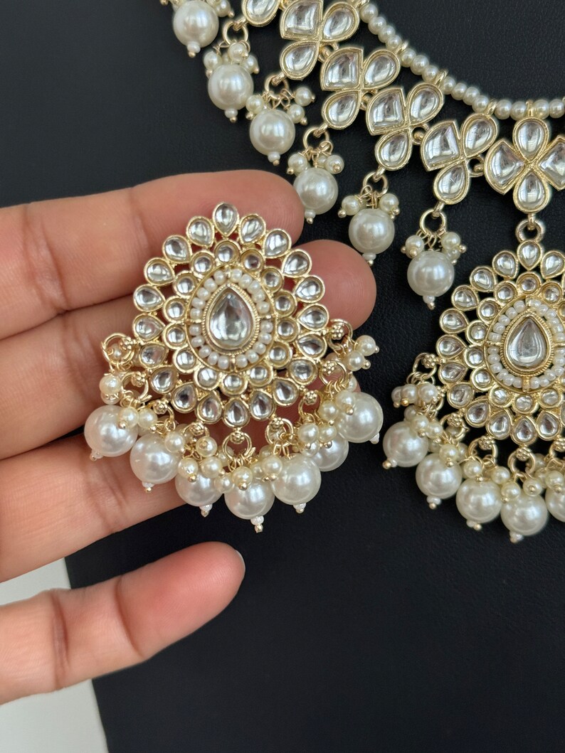 May include: A gold and white necklace and earrings set with pearl accents. The necklace features a layered design with multiple tiers of pearls and floral-shaped embellishments. The earrings are similarly designed with a round, floral-shaped center piece and a dangling pearl drop.