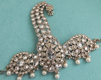 May include: An ornate, antique-style headpiece with clear, sparkling gemstones and faux pearls. The headpiece has a curved, paisley-like design with a central teardrop-shaped gem. The piece is set against a teal background.