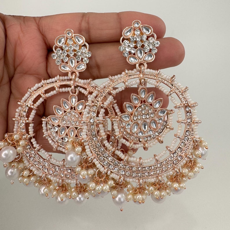 May include: A pair of large, round, gold-tone earrings with white pearl accents. The earrings feature intricate designs with multiple layers of sparkling rhinestones.