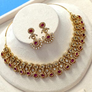 May include: A gold-toned necklace and matching pair of drop earrings featuring a repeating teardrop motif. Each piece is set with deep red oval stones and surrounded by small, clear, sparkling crystals arranged in a delicate, ornate pattern.