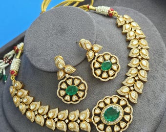 May include: A gold-toned necklace and earring set featuring oval-shaped green stones surrounded by clear, teardrop-shaped crystals. The necklace has a central floral-inspired pendant and leaf-like links, complemented by matching stud earrings.