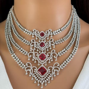 以下が含まれることがあります： 小さなキュービックジルコニアの石が4列に並んだシルバーネックレス。ネックレスには、小さなキュービックジルコニアの石で囲まれた大きな赤い正方形の石が3つ付いたペンダントが付いています。