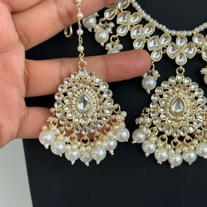 May include: A gold and white bridal necklace and earring set with pearl accents. The necklace has a layered design with multiple strands of pearls and intricate floral motifs. The earrings are teardrop shaped with a similar floral design and pearl accents.
