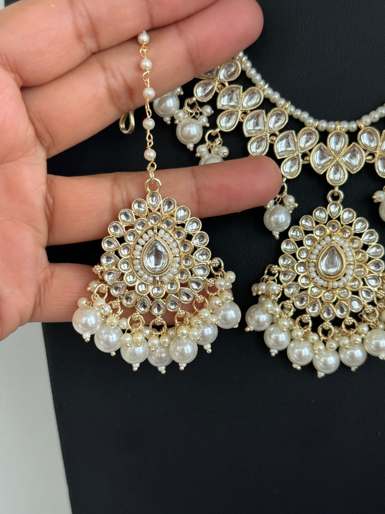 May include: A gold and white bridal necklace and earring set with pearl accents. The necklace has a layered design with multiple strands of pearls and intricate floral motifs. The earrings are teardrop shaped with a similar floral design and pearl accents.