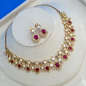 May include: A gold-toned necklace and matching earring set featuring teardrop-shaped red stones and clear round stones. The jewelry pieces are accented with small white crystals set in a metal frame, creating a detailed floral-inspired pattern.