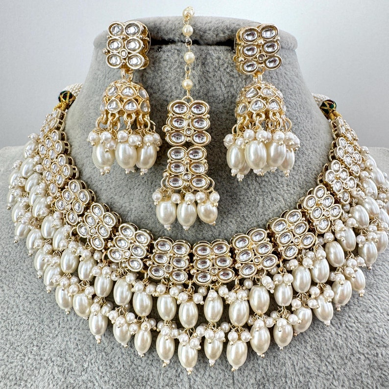 May include: A gold and white Indian bridal necklace set with pearl accents. The necklace has multiple layers of pearls and a central design of gold and clear stones. The earrings are also gold and white with a similar design to the necklace.