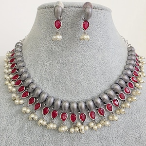 May include: A silver-coloured necklace and earring set. The necklace features textured, barrel-shaped elements with teardrop-shaped red stones and small white pearls. The matching earrings have a similar design.