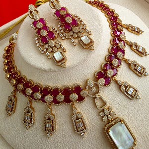 May include: A gold-toned necklace and matching earring set featuring oval-cut deep pink stones and clear pavé crystals. The necklace includes dangling rectangular pendants with white rectangular stones and a large central white rectangular stone.
