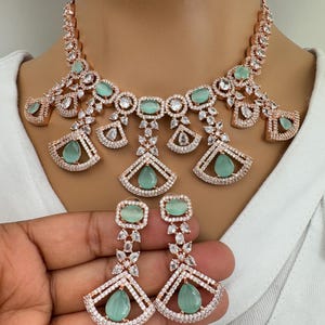 Pode incluir: Um conjunto de colar e brincos de ouro rosa com pedras transparentes e verde claro. O colar tem um design em forma de leque com v&aacute;rios n&iacute;veis. Os brincos s&atilde;o longos e pendentes com um design semelhante em forma de leque.