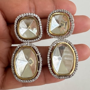 May include: Four clear, faceted gemstone earrings. Two pairs of earrings are visible, each with a gold-colored inner frame, a silver-colored outer frame, and small, clear accent stones. The earrings are held in a person's hand.