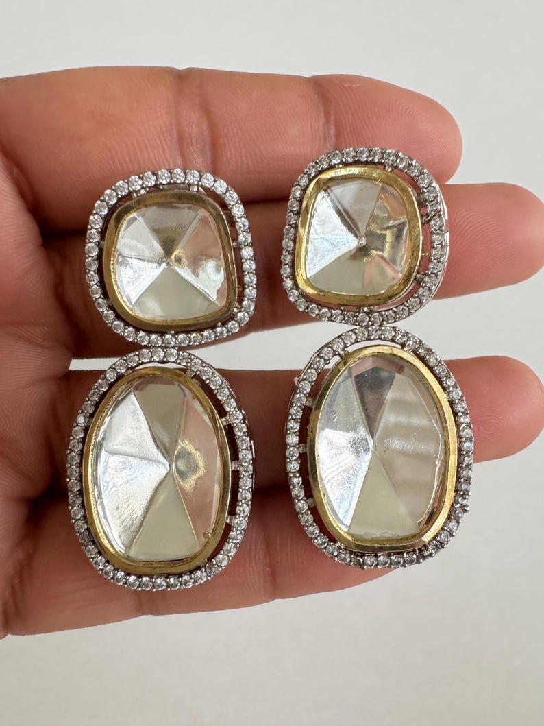 May include: Four clear, faceted gemstone earrings. Two pairs of earrings are visible, each with a gold-colored inner frame, a silver-colored outer frame, and small, clear accent stones. The earrings are held in a person's hand.