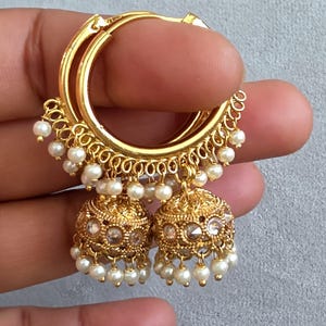May include: Gold-toned hoop earrings with dangling pearl accents and ornate, bell-shaped dangles. The earrings feature a detailed filigree design and small, clear gemstones. The hoops are held by a hand against a neutral background.