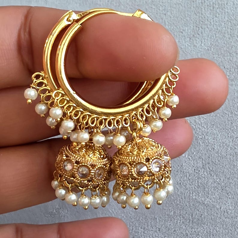 May include: Gold-toned hoop earrings with dangling pearl accents and ornate, bell-shaped dangles. The earrings feature a detailed filigree design and small, clear gemstones. The hoops are held by a hand against a neutral background.