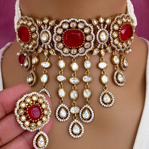 May include: A gold-toned choker necklace with a central red gemstone and dangling white teardrop-shaped accents. The necklace features a floral design with clear, round gemstones. A matching pendant is held in the hand.