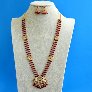Pode incluir: Um colar comprido com dois fios de contas vermelho-escuras, espaçadores dourados e um pendente central com pedras vermelhas e verdes, acompanhado por um par de brincos jhumka em forma de sino com adornos de contas vermelhas.