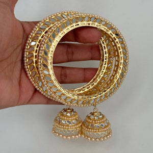 Könnte beinhalten: Goldfarbene Armreifen mit Spiegel- und Perlenverzierungen. Die Armreifen sind gestapelt, mit glockenförmigen Anhängern. Die Anhänger sind ebenfalls mit Spiegeln und Perlen besetzt, was einen glamourösen Look erzeugt.