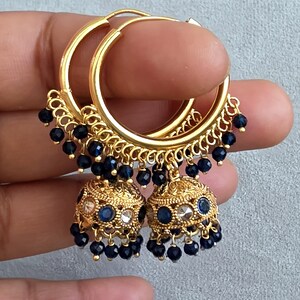 May include: Gold-toned hoop earrings with intricate filigree detailing. Each earring features a bell-shaped charm adorned with dark blue and clear gemstones, and dangling dark blue beads. The earrings are held up by a hand.