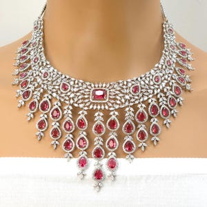 Puede incluir: Un deslumbrante collar plateado con una gran piedra central rectangular de color rojo rubí. El collar está adornado con numerosos cristales claros y brillantes y piedras en forma de lágrima de color rojo rubí, creando un diseño lujoso y elegante.