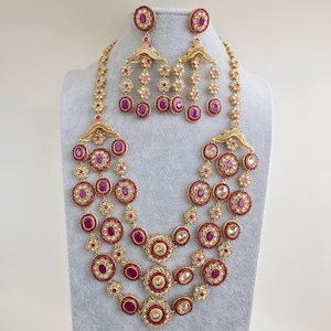 Puede incluir: Un conjunto de collar y pendientes dorados. El collar presenta múltiples capas de piedras circulares de color rubí y pequeños detalles florales. Los pendientes tienen un diseño de araña con piedras de rubí a juego. La joyería se exhibe en un maniquí gris.